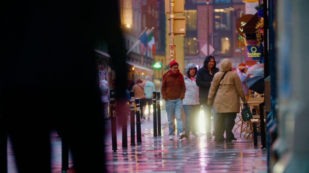 Nighttime telephoto image of pedestrians on rain-slicked Dublin streets with city lights reflecting on pavement