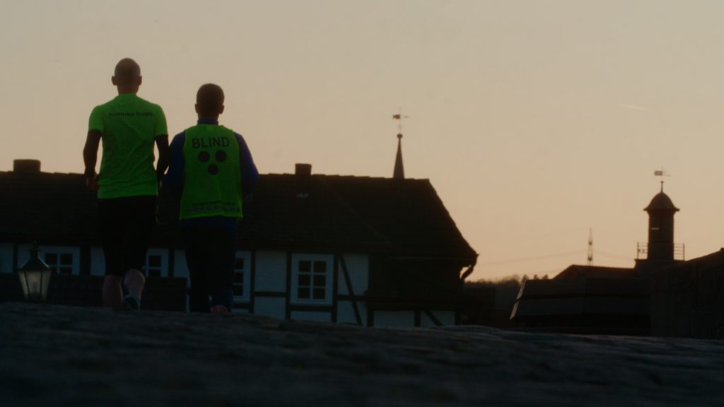 Backlit telephoto image of blind runner and guide silhouetted against setting sun