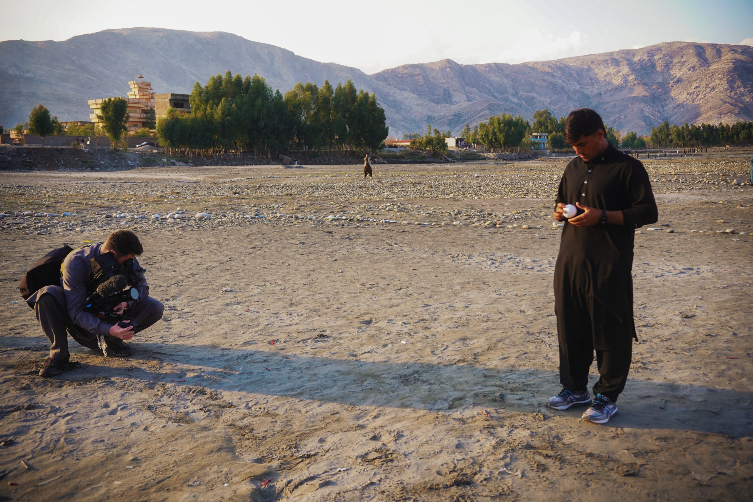 Killian Bayer capturing a low-angle shot of the Afghanistan documentary protagonist bowling a cricket ball on a riverbank, framed by a mountain range in the background during sunset.