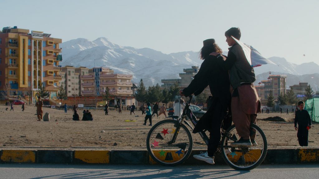 Two boys on a bicycle watching a cricket match in front of mountains in Kabul