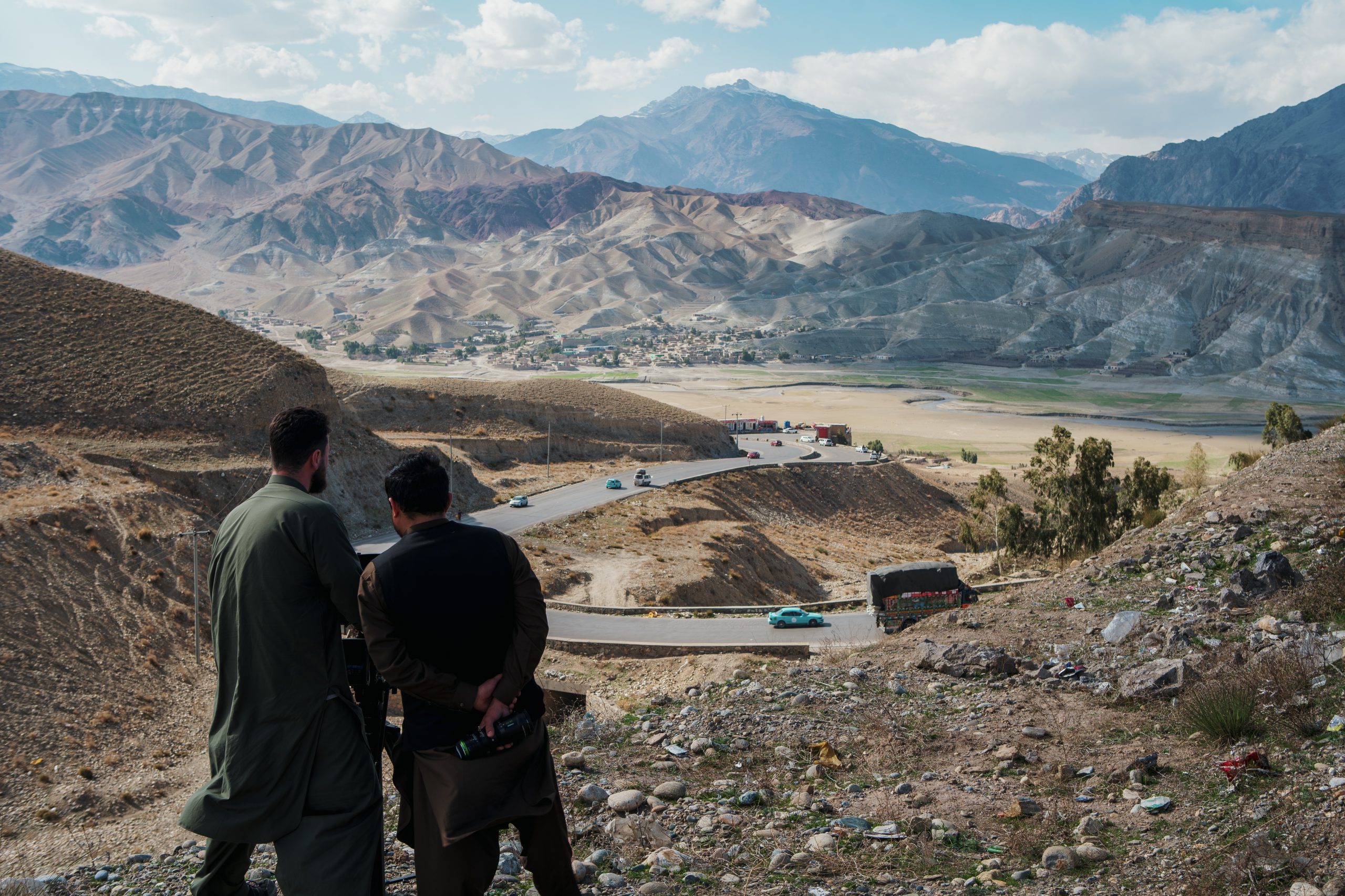 Killian Bayer and his colleague capturing landscape shots in the mountains of Afghanistan, while trucks and traffic struggle up a winding mountain road in the background.