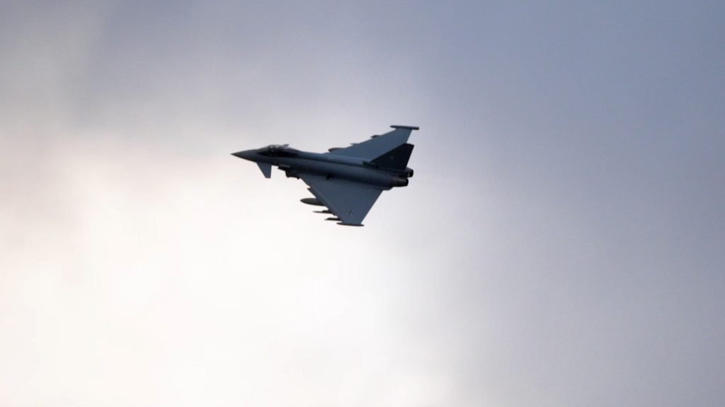Silhouetted Eurofighter jet banking sharply against a backdrop of bright clouds after takeoff